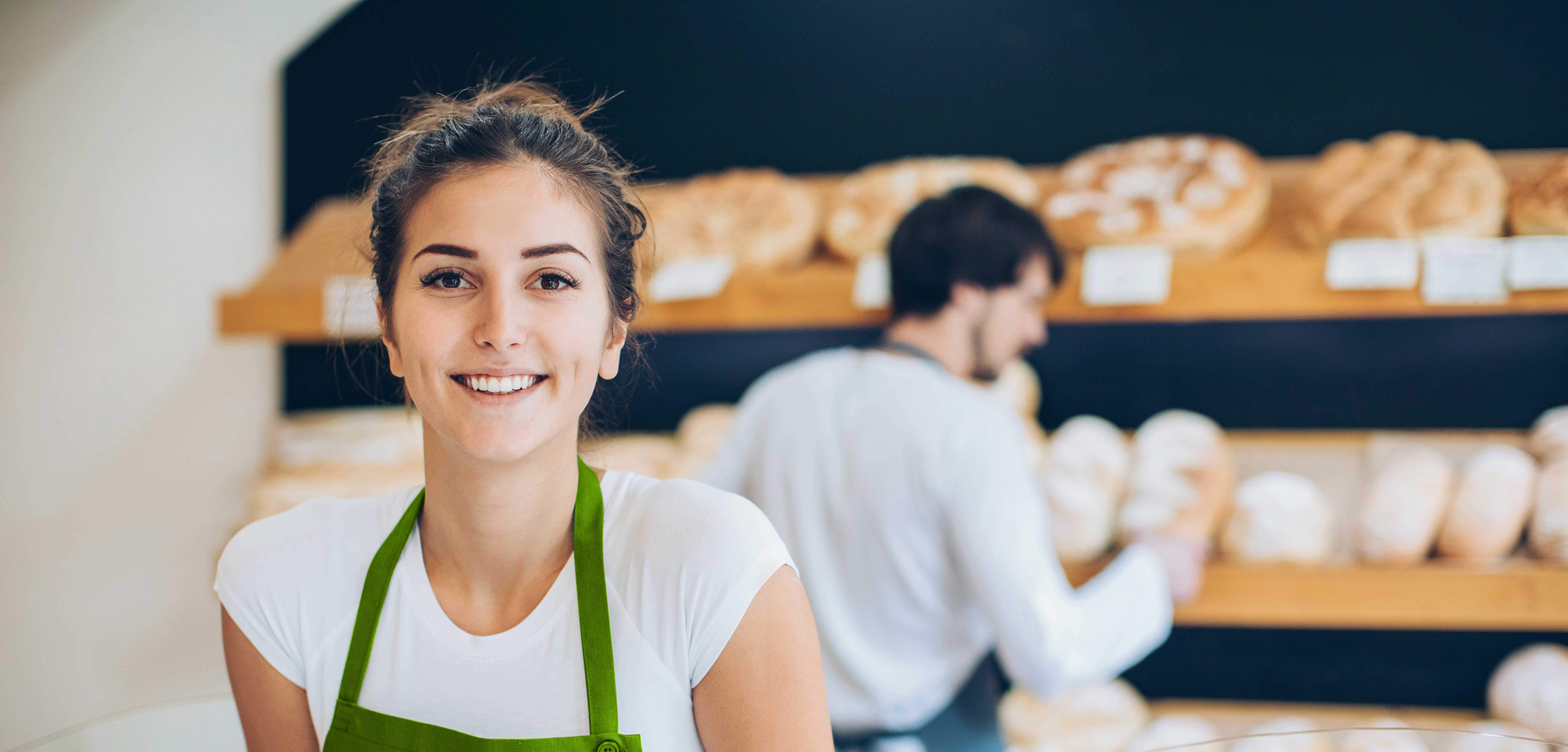 Verkäuferin einer Bäckerei mit zweitem Verkäufer und Broten im Hintergrund