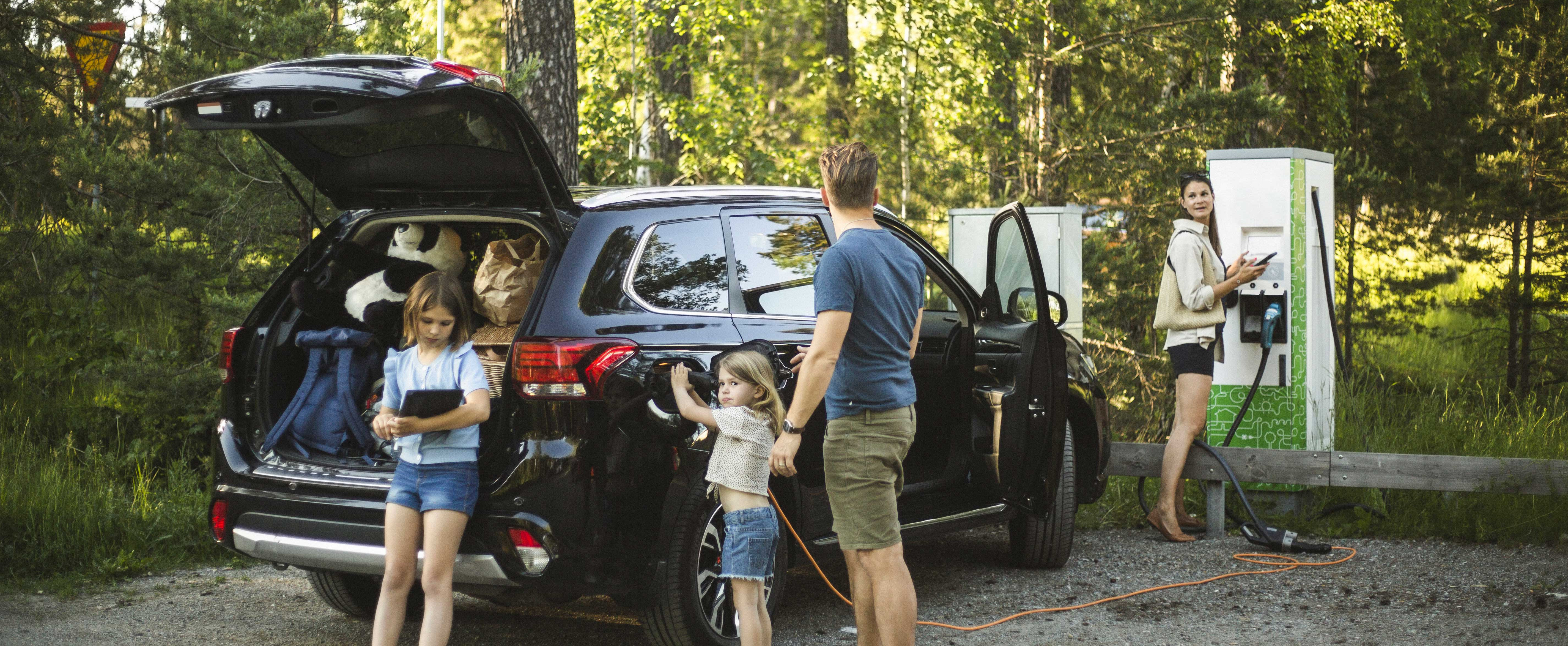 Eine Familie mit vollgepacktem Auto steht auf einem Parkplatz im Wald und lädt ihr E-Auto.